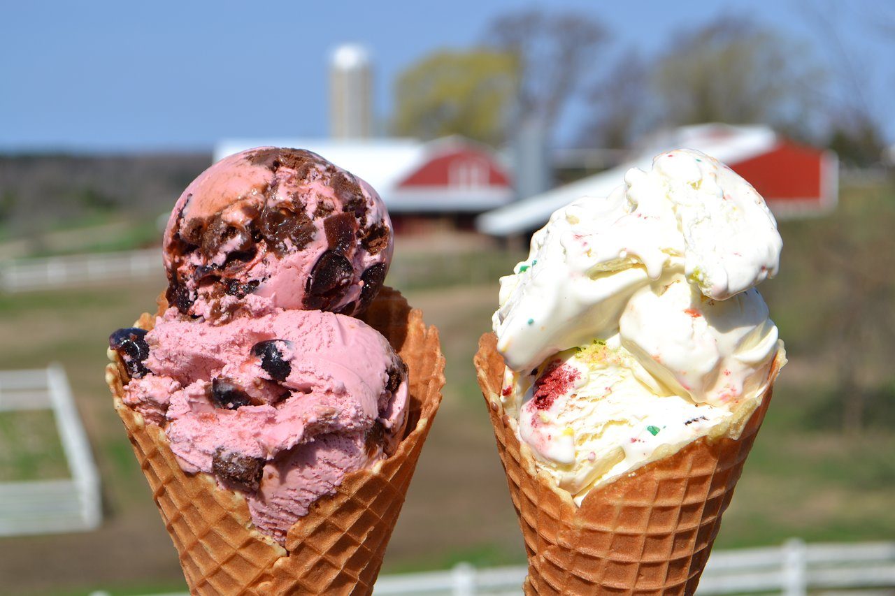 Two ice cream cones held upright against a blurred rural background of red barns and green fields.