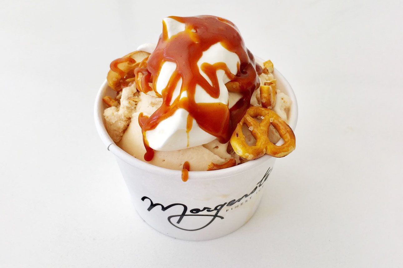 Ice cream topped with caramel and pretzels sits in a white cup on a plain surface. The cup displays "Morgenstern's" in elegant script.