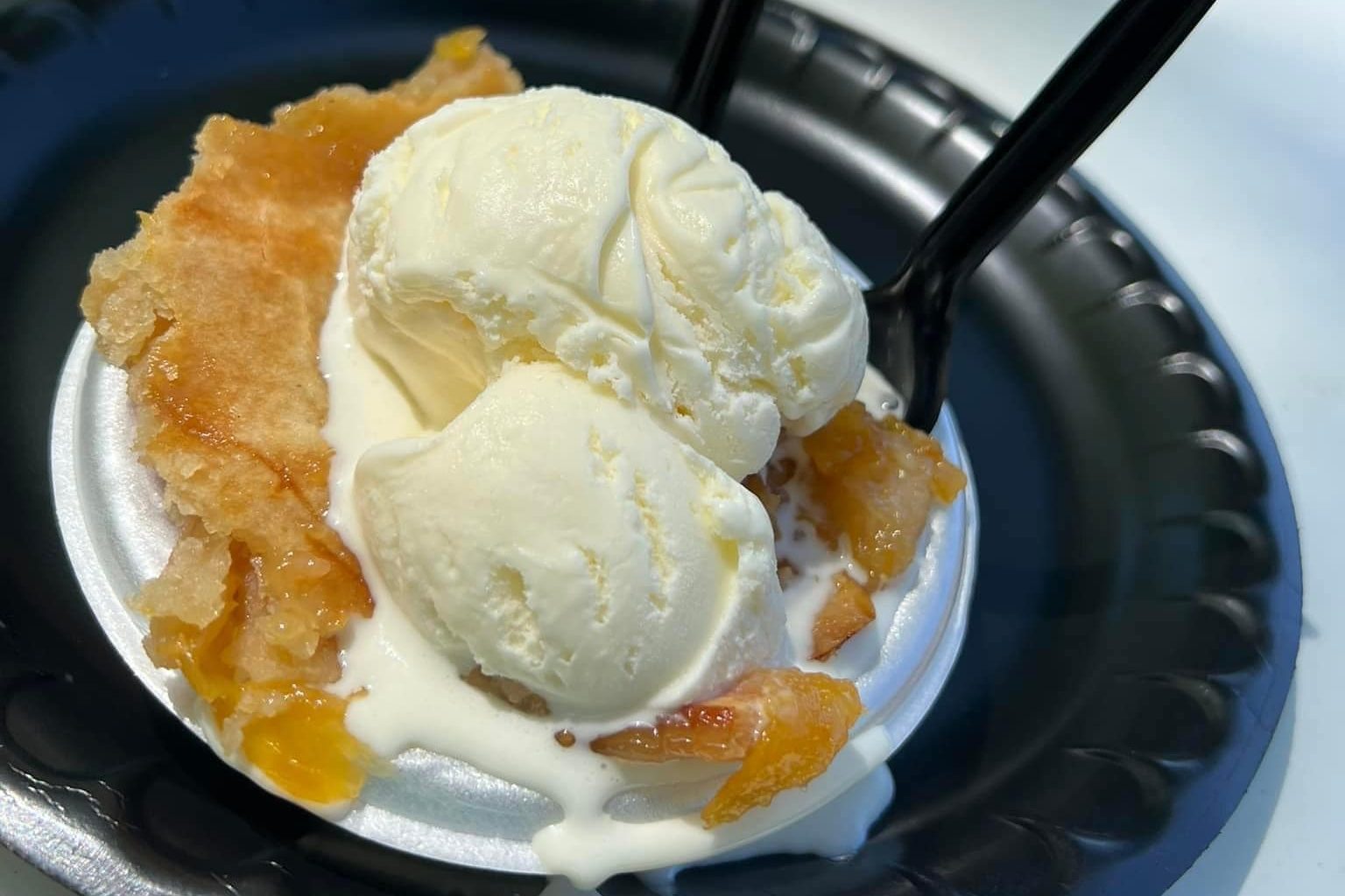 Ice cream melts over peach pie slice on a black plastic plate, with two black forks beside it in a bright setting.