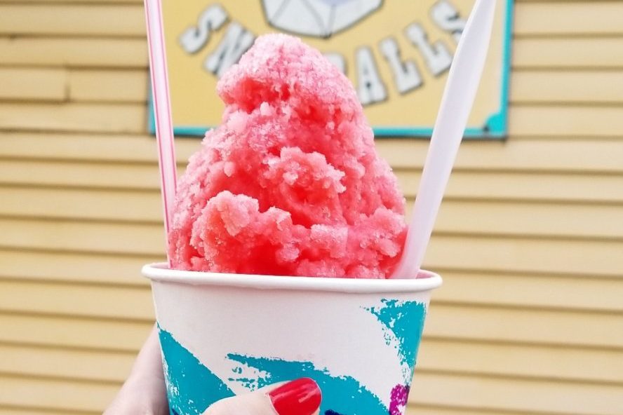 A hand holds a pink shaved ice dessert in a colorful cup, positioned in front of a wooden building.