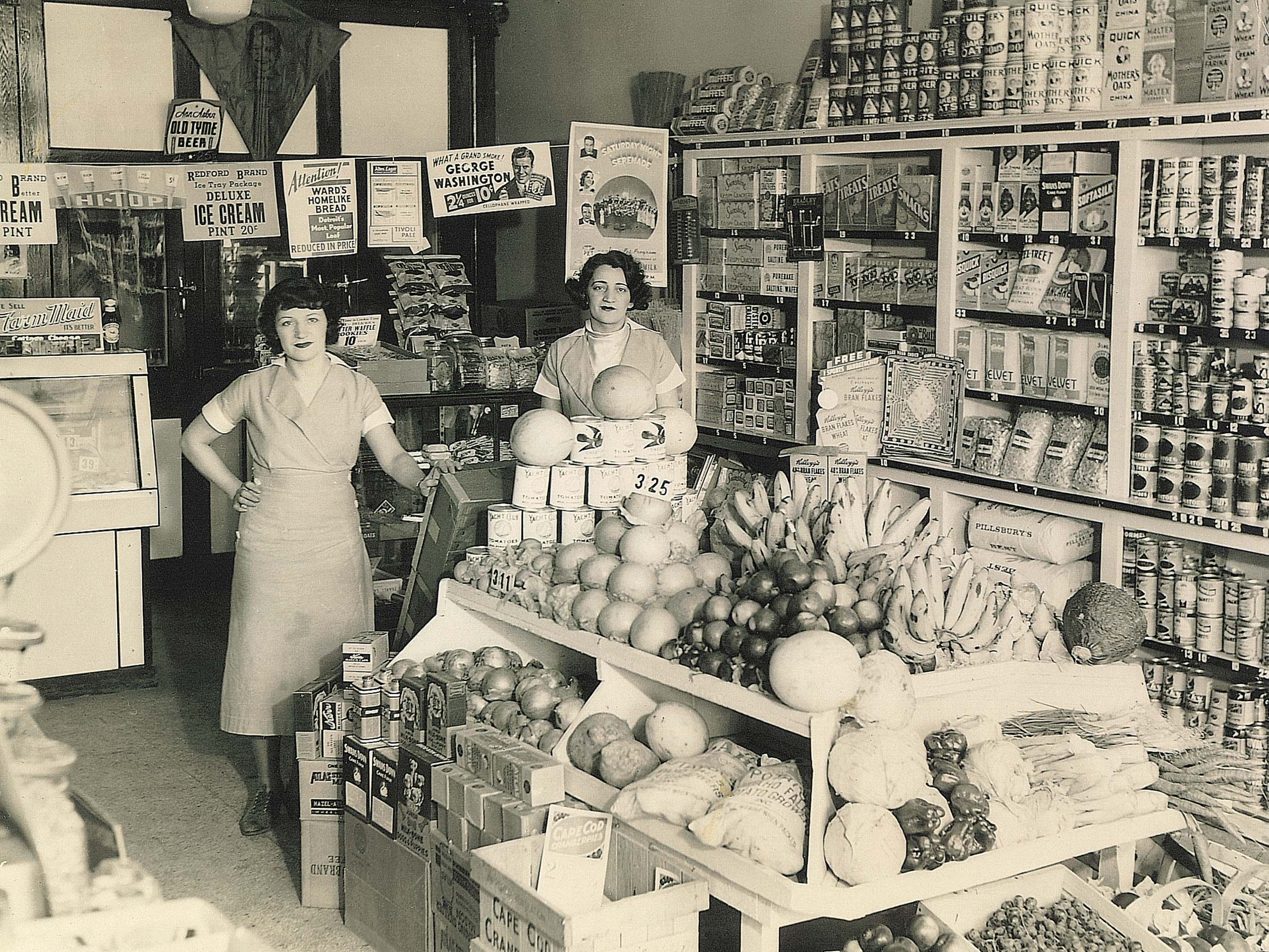 Two women stand behind a counter displaying fruits and vegetables in a vintage grocery store, surrounded by shelves of canned goods and advertisements.