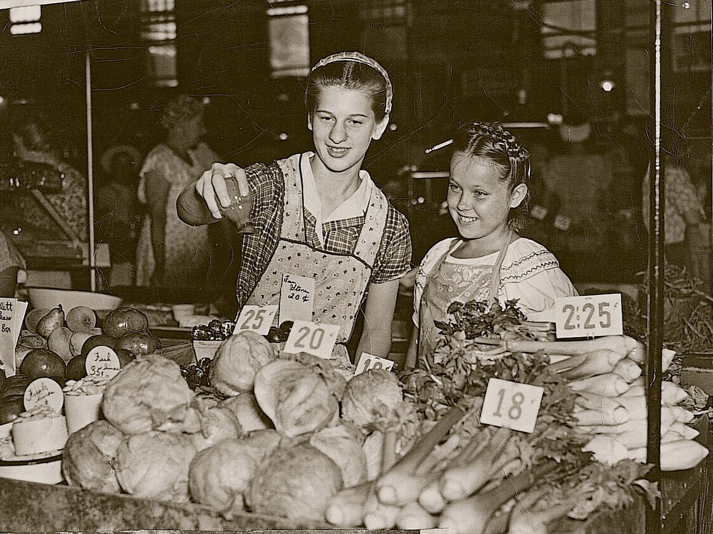 Two girls examine produce at a market stall, surrounded by vegetables with price tags.