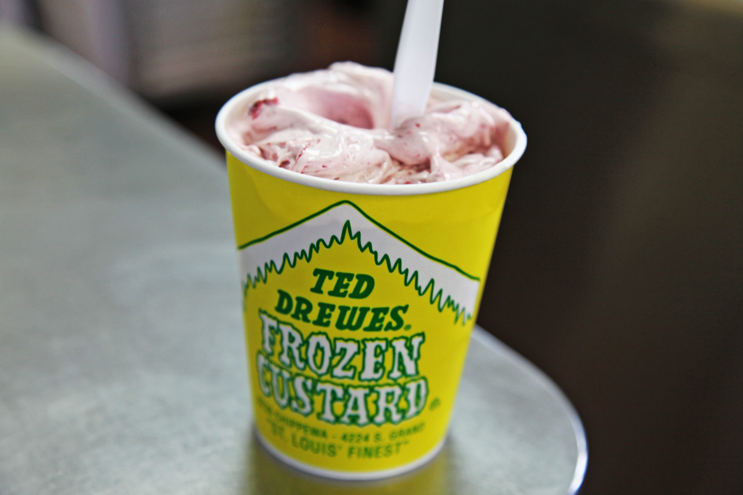 Frozen custard in a yellow cup with green text, sitting on a metallic surface, with a white spoon inserted.