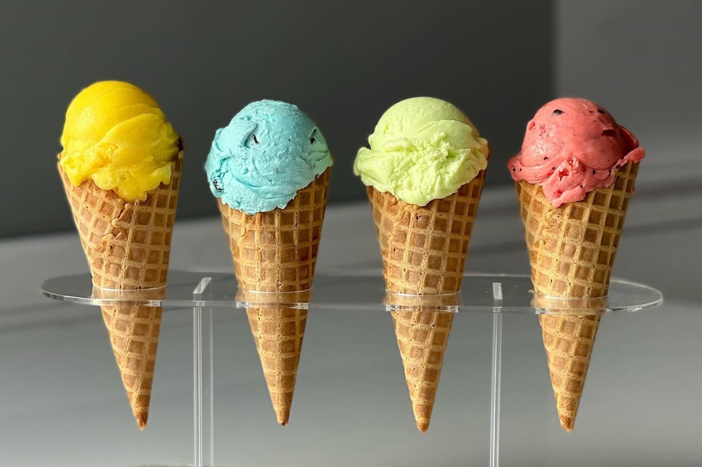 Four ice cream cones, vibrant colors, stand upright on a transparent holder against a neutral background.