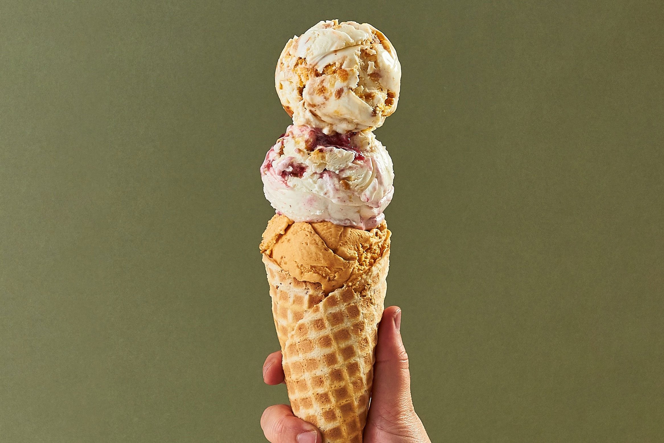 A hand holds a waffle cone with three scoops of ice cream against a plain green background.