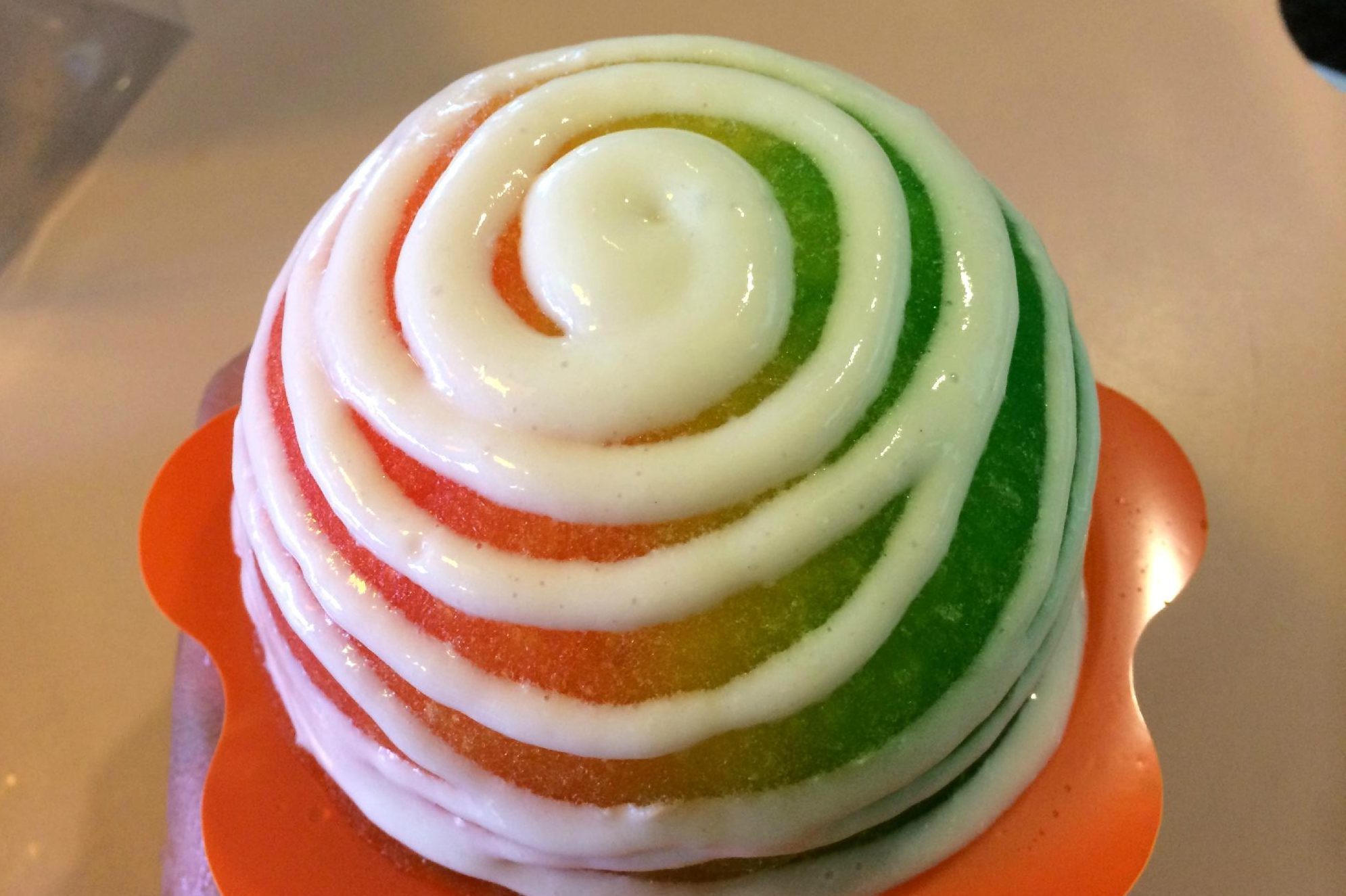 Colorful shaved ice swirled with white cream, resting on an orange holder, placed on a light surface.