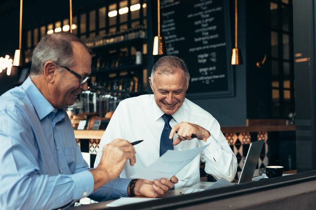 Two men, smiling, review documents in a cozy cafe setting with warm lighting and a menu board in the background.