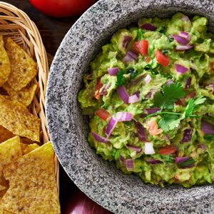Guacamole sits in a stone bowl, garnished with cilantro and diced onions, surrounded by a basket of tortilla chips and fresh vegetables.