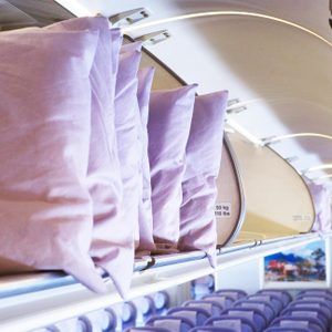 Pillows stacked inside an open airplane overhead compartment; the cabin features rows of purple seats underneath.
