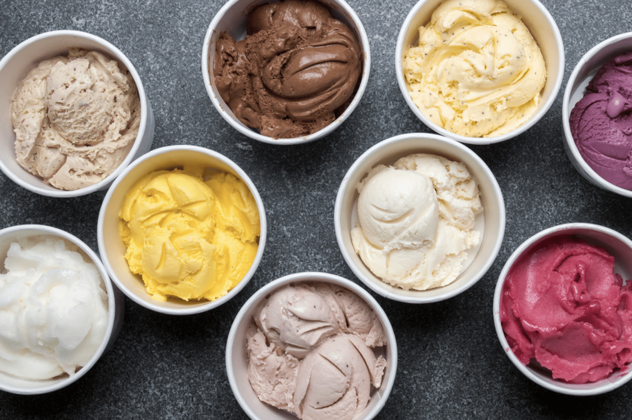 Bowls hold colorful ice cream scoops, arranged closely on a dark stone surface.