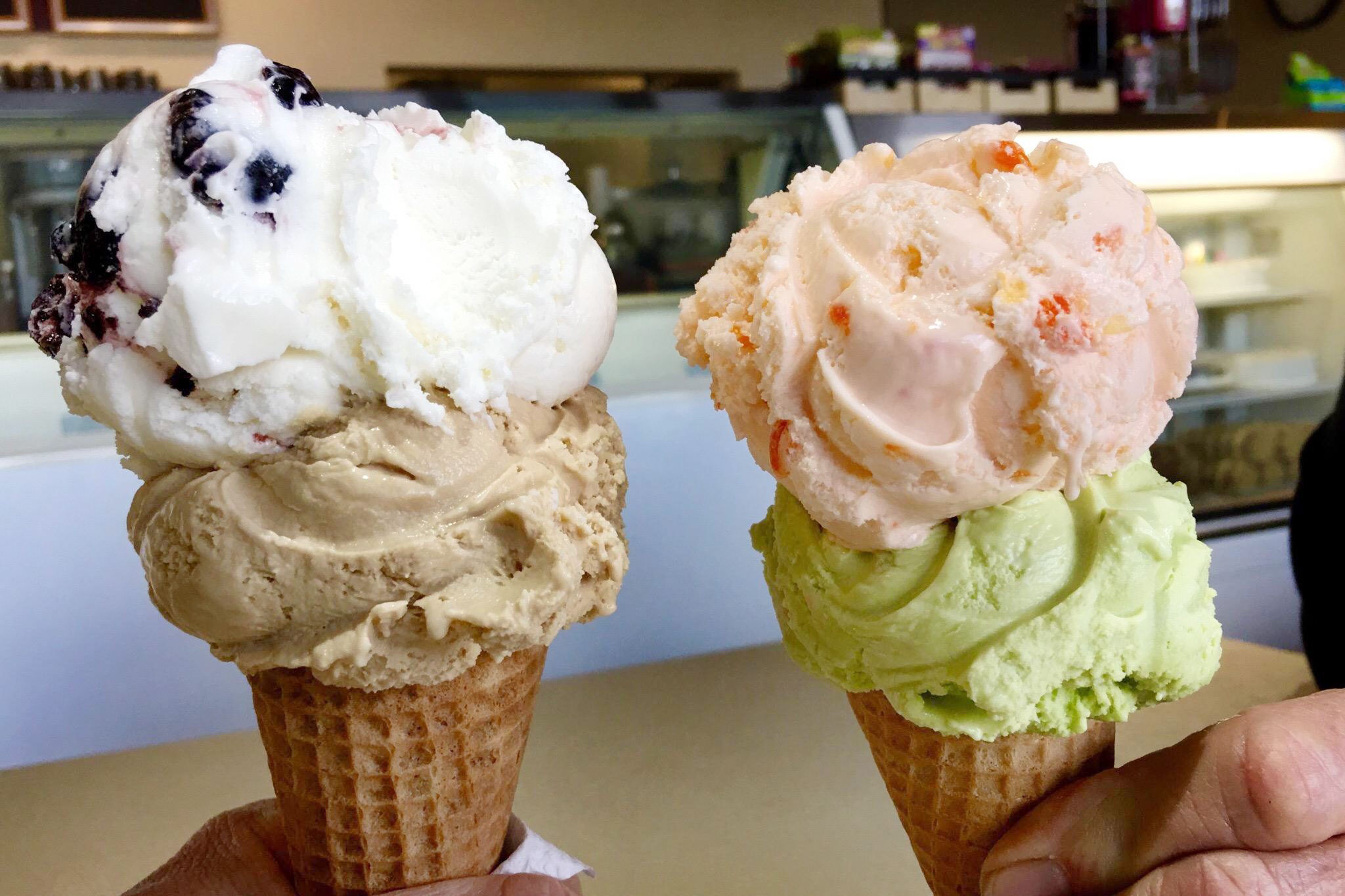 Two hands hold ice cream cones with multiple scoops, creamy textures and different flavors, inside an ice cream shop with a counter and shelves in background.