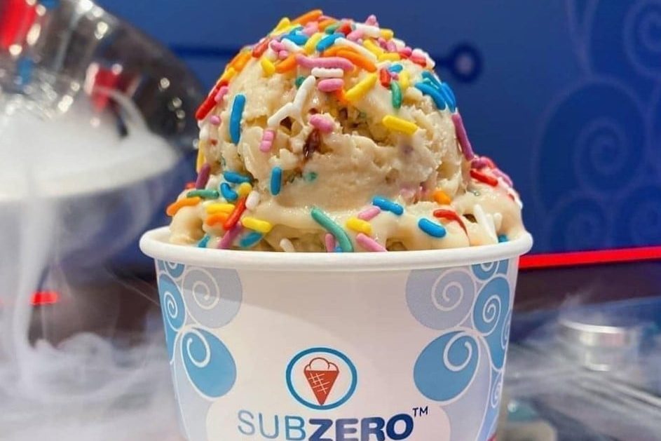 Ice cream sits in a cup, topped with colorful sprinkles. The cup features the logo "SUBZEROâ„¢." Whipped cream and bowls are in the background.