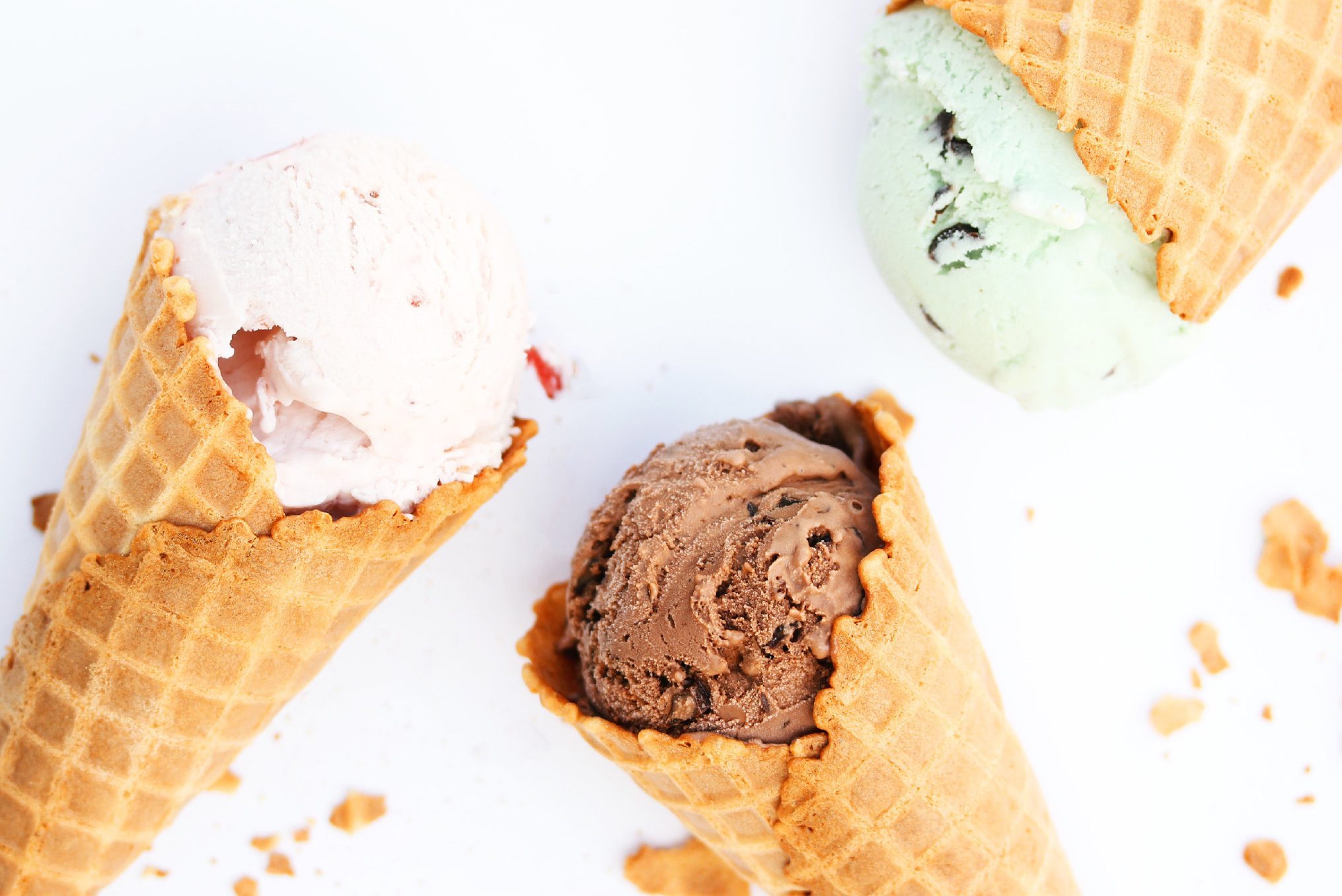 Three waffle cones hold scoops of strawberry, chocolate, and mint ice cream on a white background with scattered crumbs.
