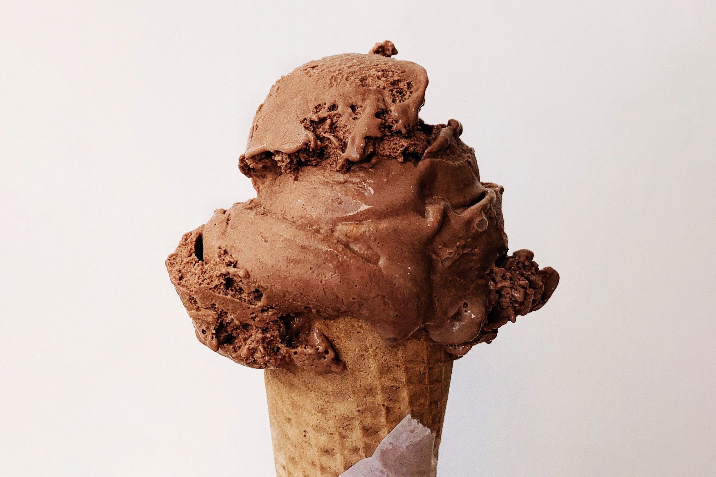 Chocolate ice cream sits atop a waffle cone, melting slightly against a plain, light background.