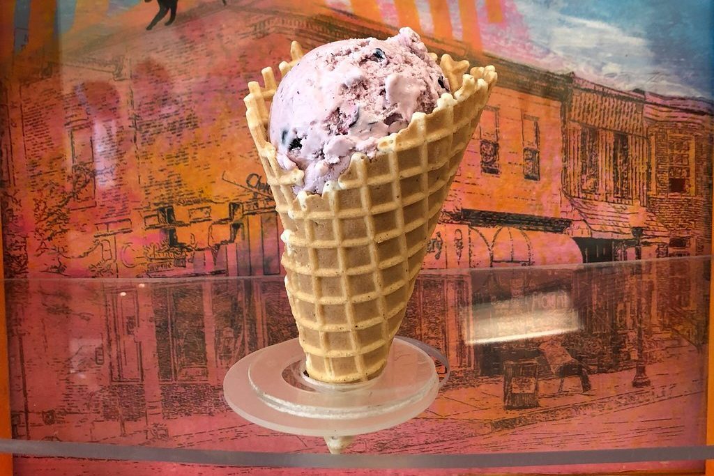 Waffle cone holds purple ice cream; positioned upright in stand against a colorful, vintage-style mural background depicting a street scene.