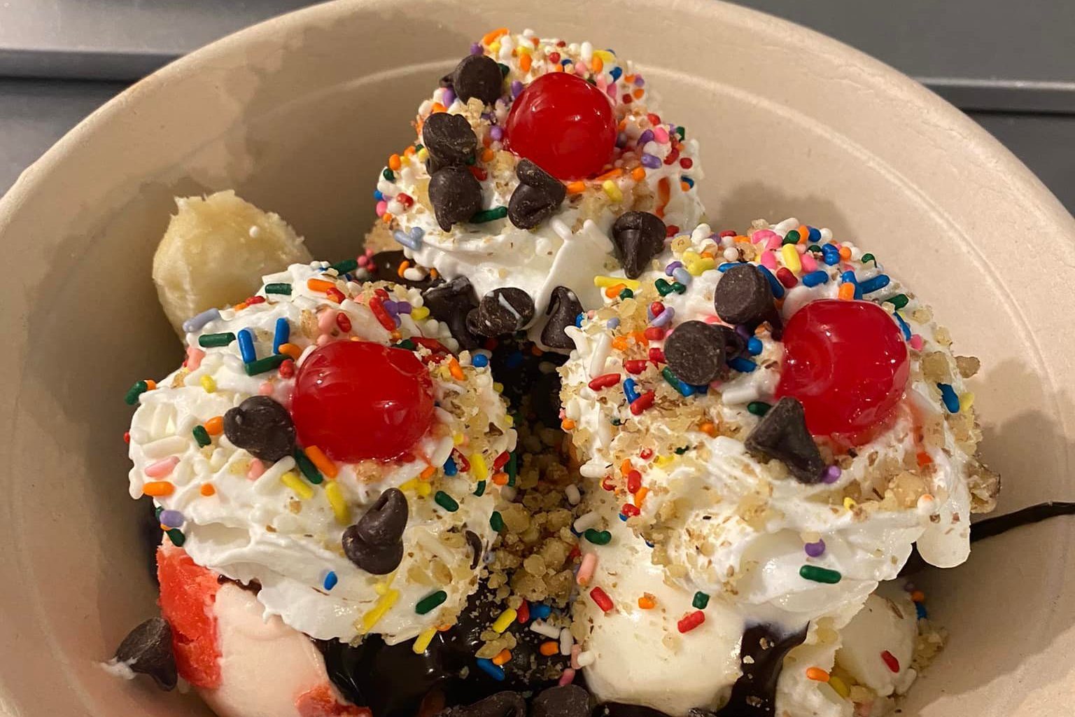 Banana split topped with whipped cream, cherries, chocolate chips, and colorful sprinkles, served in a beige bowl.