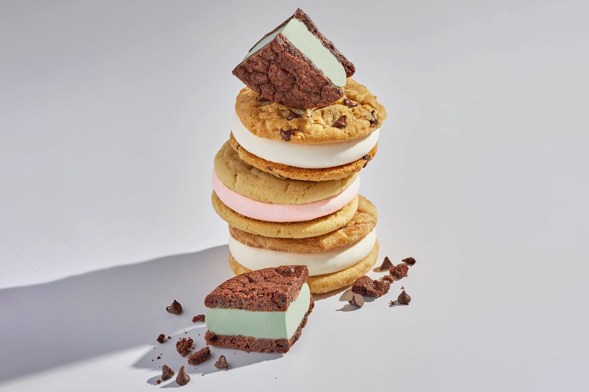 Ice cream sandwiches stacked, topped with a halved brownie-ice cream piece, surrounded by scattered chocolate chips on a light surface.