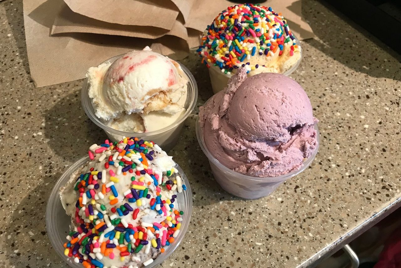Four cups of colorful ice cream sit on a speckled countertop, topped with rainbow sprinkles, against a backdrop of folded brown napkins.