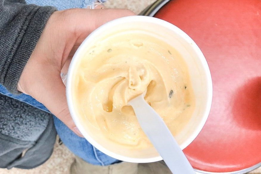 Cup of creamy orange frozen dessert, being held, with a white spoon inside, over a red chair.