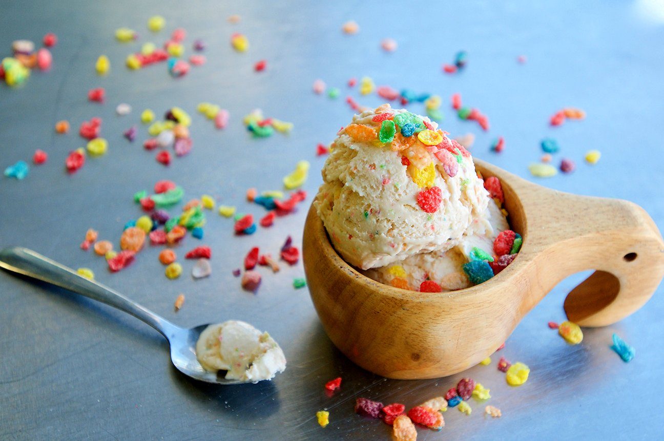 Ice cream sits in a wooden bowl, topped with colorful cereal, on a metal table with scattered cereal and a spoon holding a small scoop.