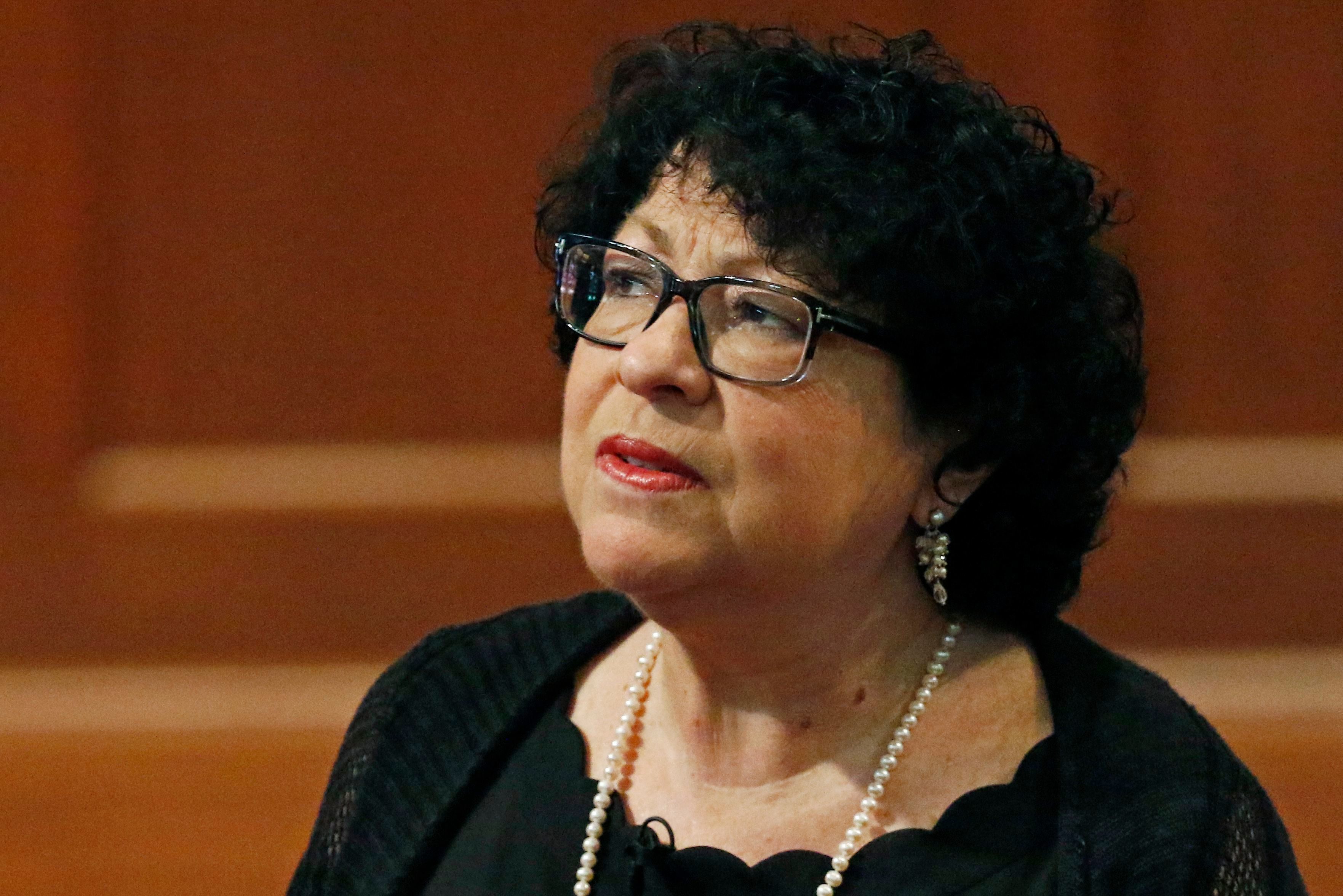 A woman with curly hair, wearing glasses and pearls, gazes upward in a well-lit indoor setting.
