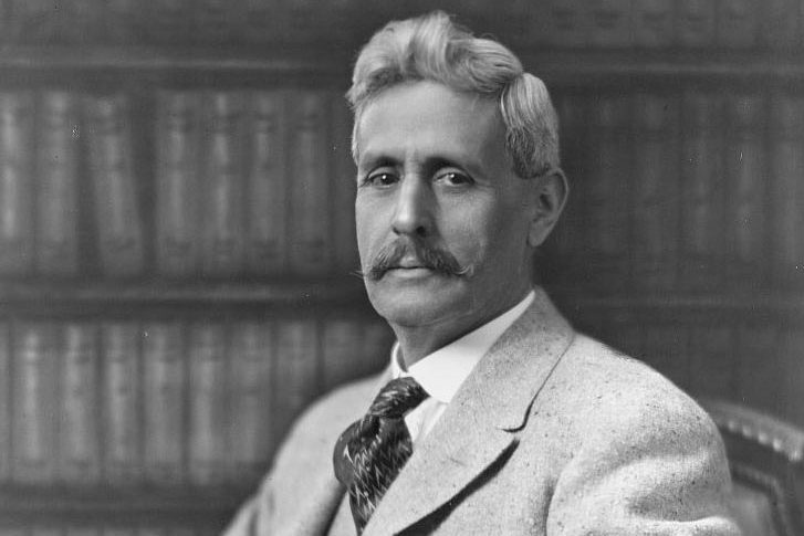 Man sitting calmly against a backdrop of shelves filled with books, wearing a suit and tie, gazing directly at the camera.