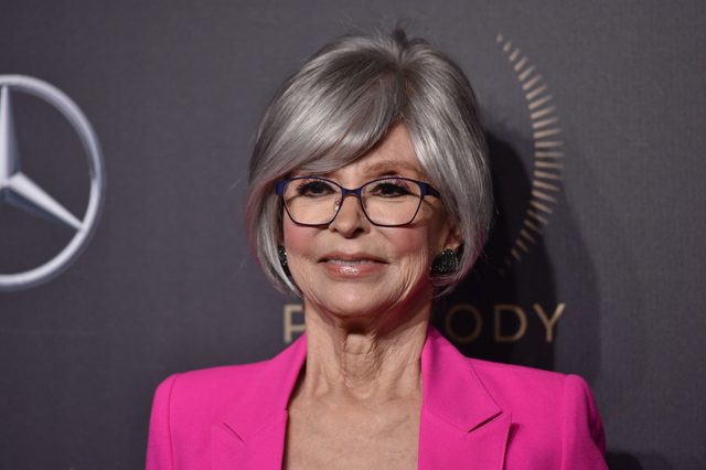 A person wearing glasses smiles, dressed in a bright pink jacket, standing against a dark background with a circular emblem and text.