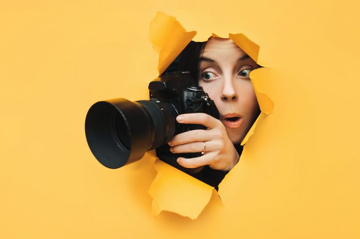 A paparazzi funny girl with a camera looks out from cover and looks at what is happening with her mouth open. Yellow paper, torn hole. Tabloid press. In search of the plot for photo stocks.