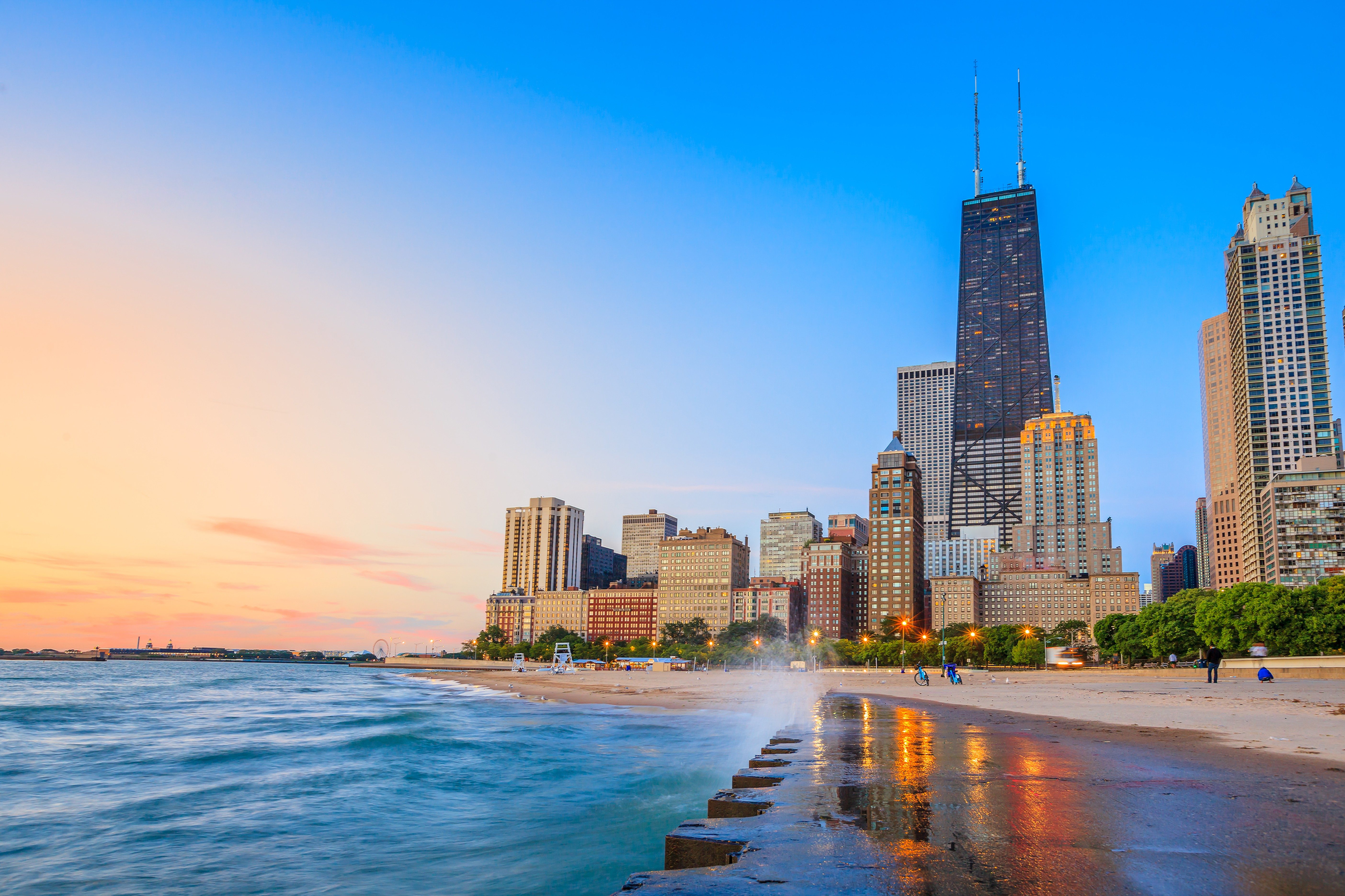 Skyscrapers stand tall against a colorful sunset, with waves crashing on the foreground shore and people walking along the sandy beach.