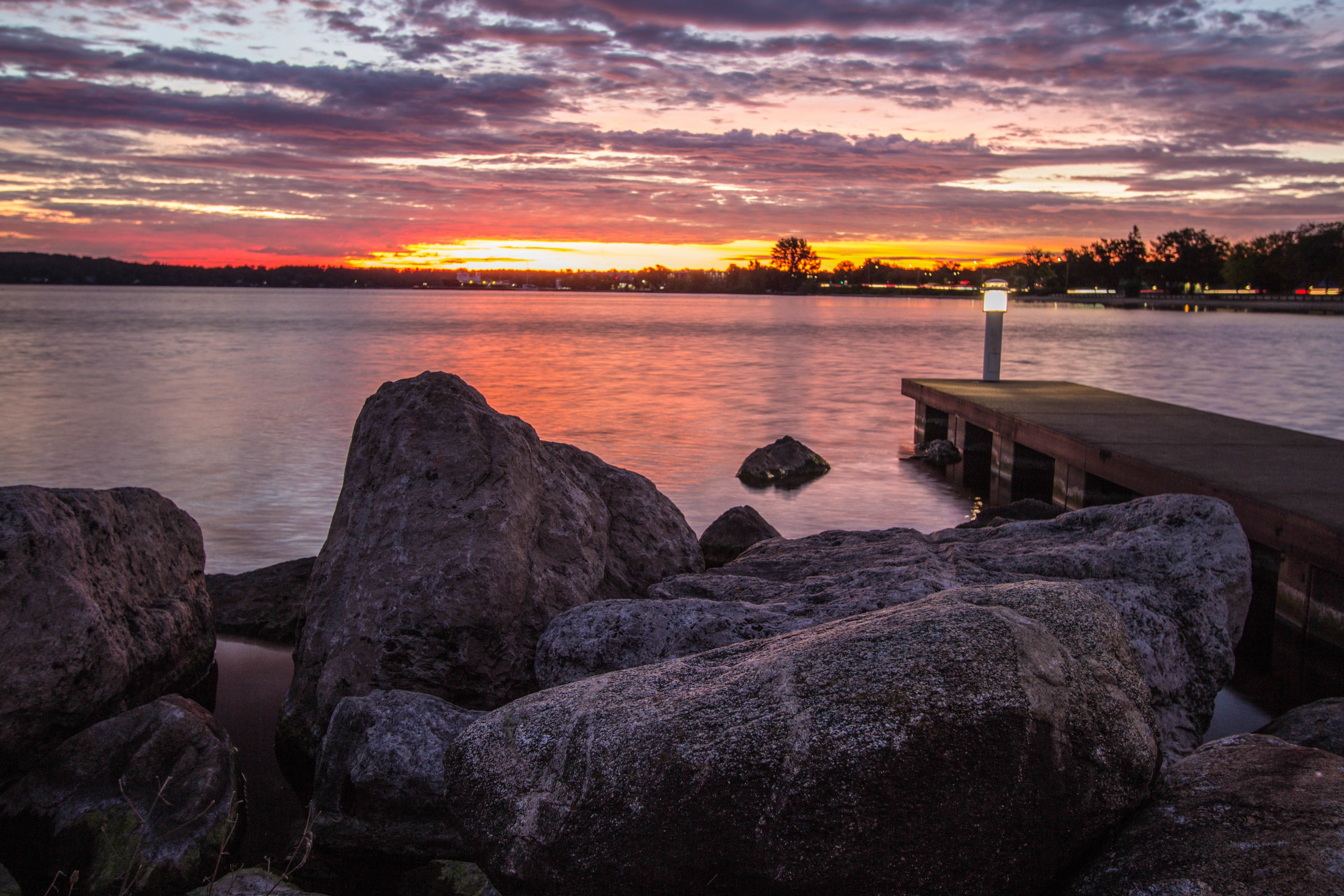 Rocks sit quietly by a calm lake at sunset, with a wooden dock and distant lit sky.