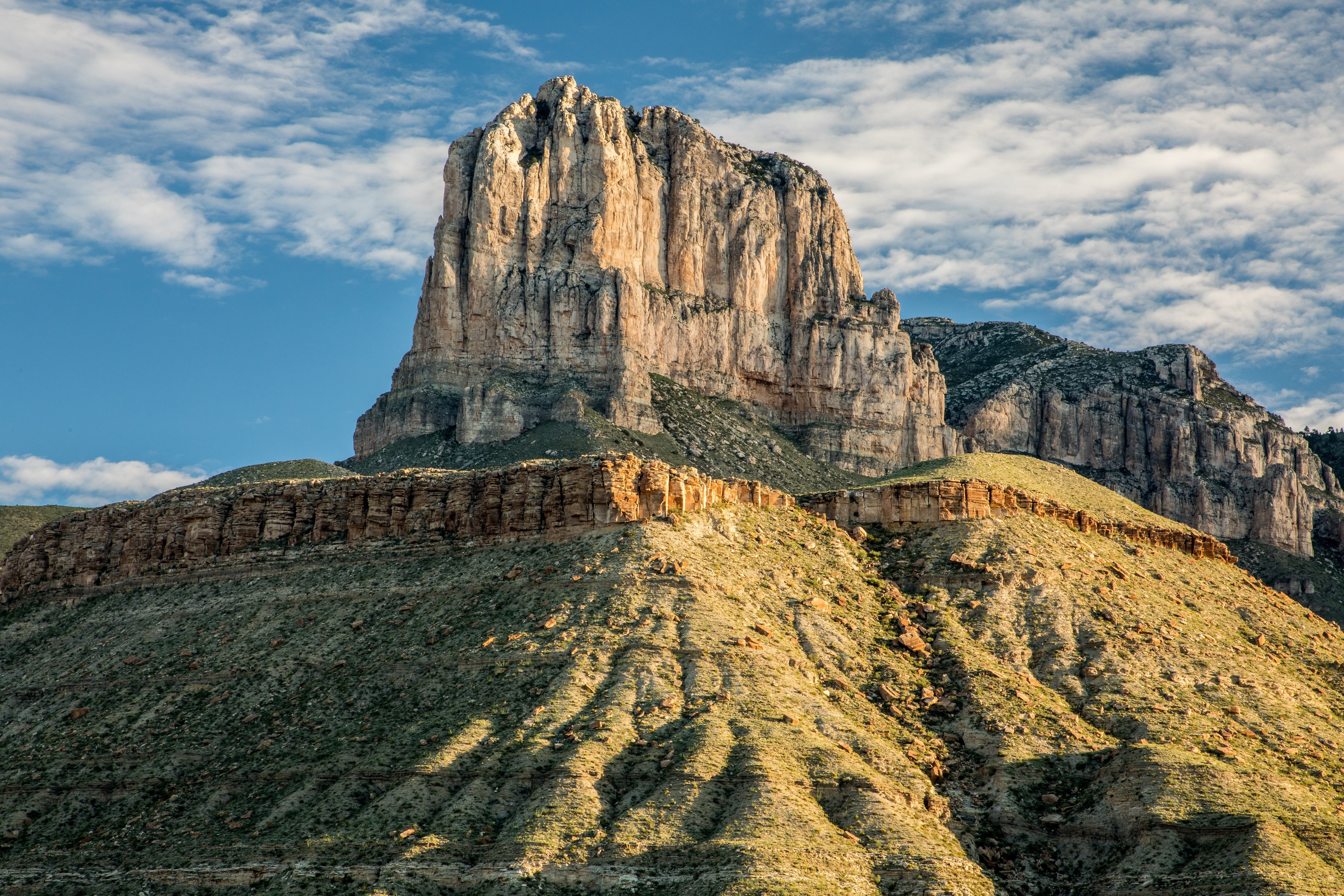Steep mountain rises dramatically, bathed in sunlight, under a partly cloudy sky, surrounded by rugged terrain and shadows.