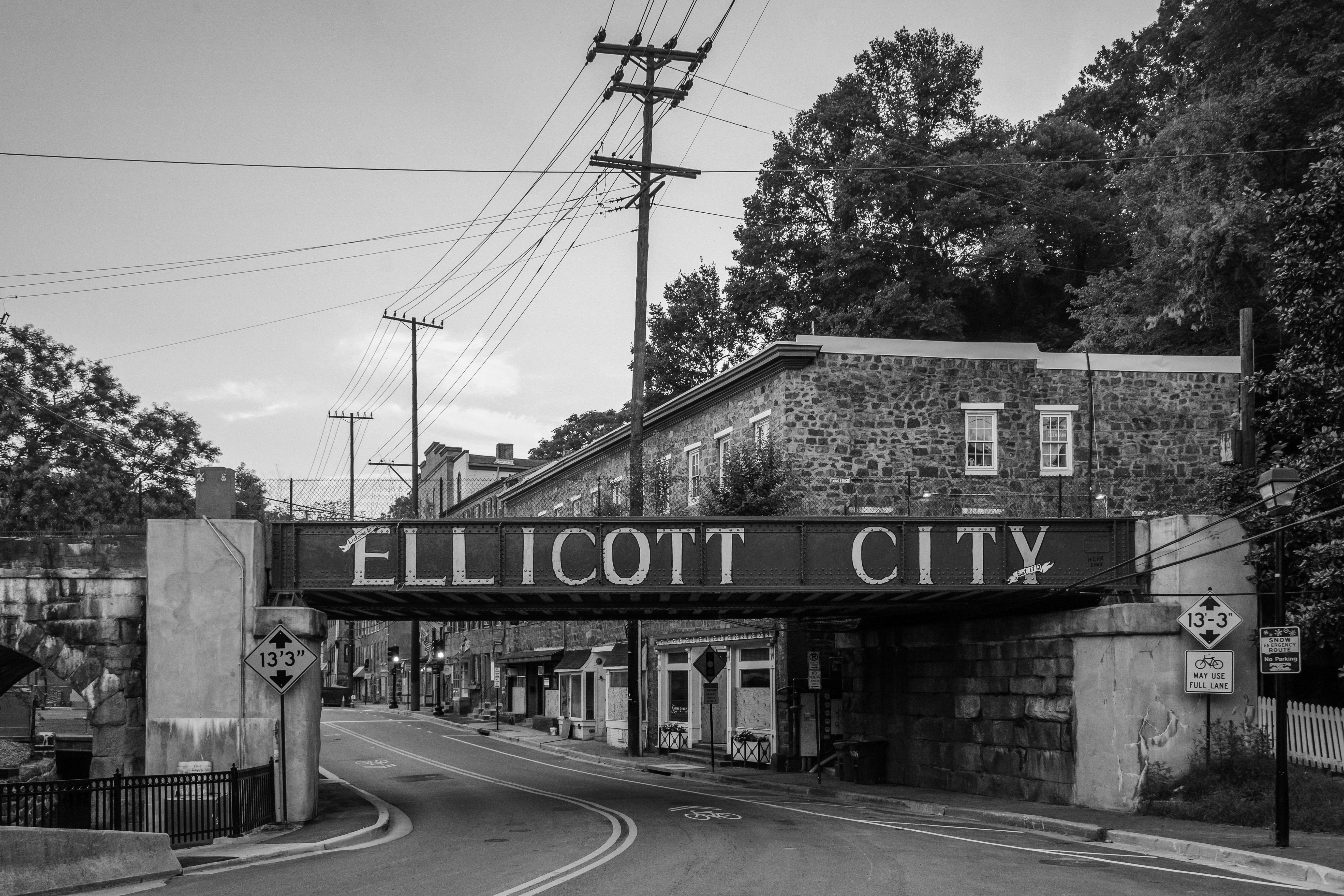 A railway bridge displays "ELLICOTT CITY" above a narrow road, surrounded by old brick buildings and trees, with utility poles and power lines overhead.
