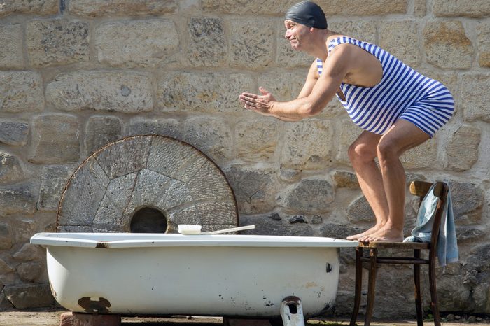 Fat man in retro swimsuit jumps to the outdoor bathtub. Funny swimmer in vintage style and bathing cap starts to swim in the tub. Home vacations. Domestic holiday.