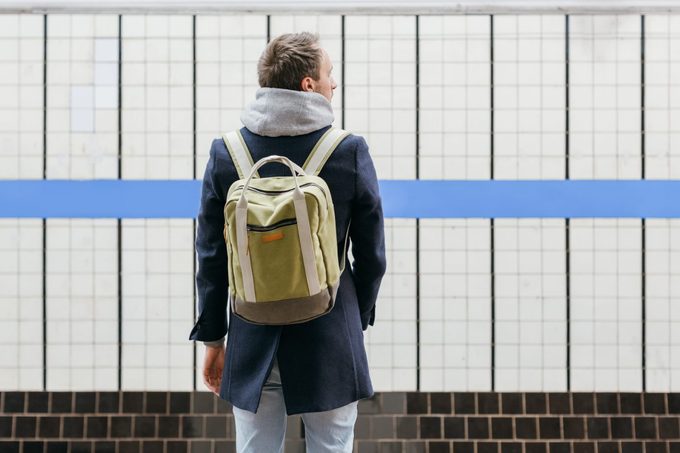 Lonely adult handsome man in a blue coat with a green backpack behind standing on the platform waiting for the train. Selective focus on male. Copy space. Morning commute. Urban life concept.