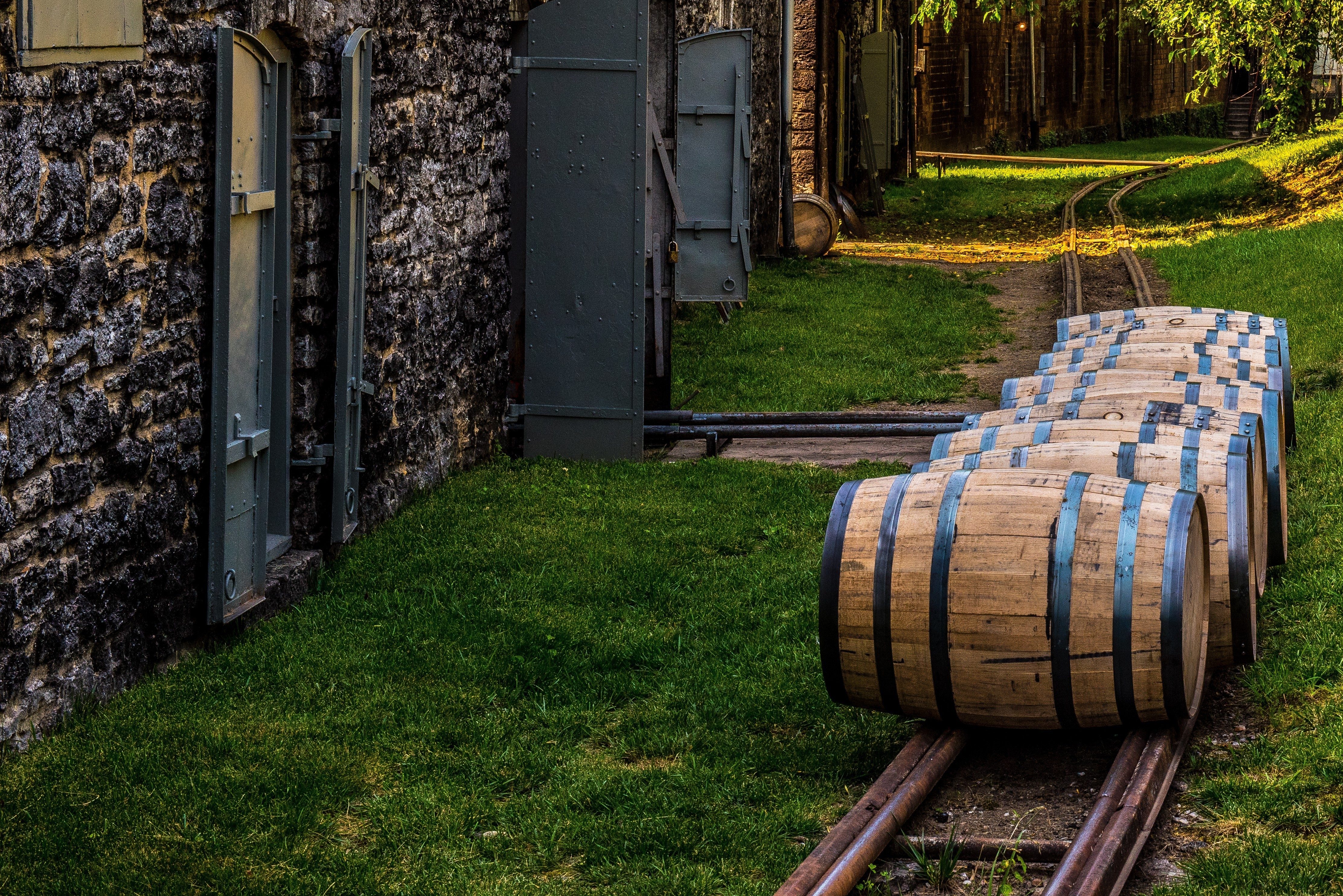 Barrels roll along narrow tracks beside a stone building, surrounded by grass and trees, in a rural outdoor setting.