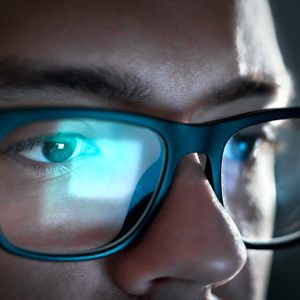 Person wearing glasses reflects glowing screen, focused in dimly lit room.