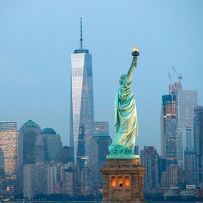 The Statue of Liberty holds a torch against a backdrop of New York City's skyline, including tall skyscrapers at dusk.