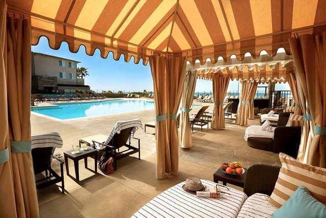 Lounge chairs rest under cabanas beside a pool, amid a resort setting with palm trees and buildings in the background.