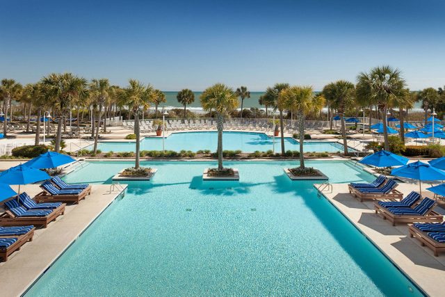 Pool, shimmering under sunlight, surrounded by palm trees and blue-striped loungers. Blue umbrellas provide shade, with the ocean visible in the background.