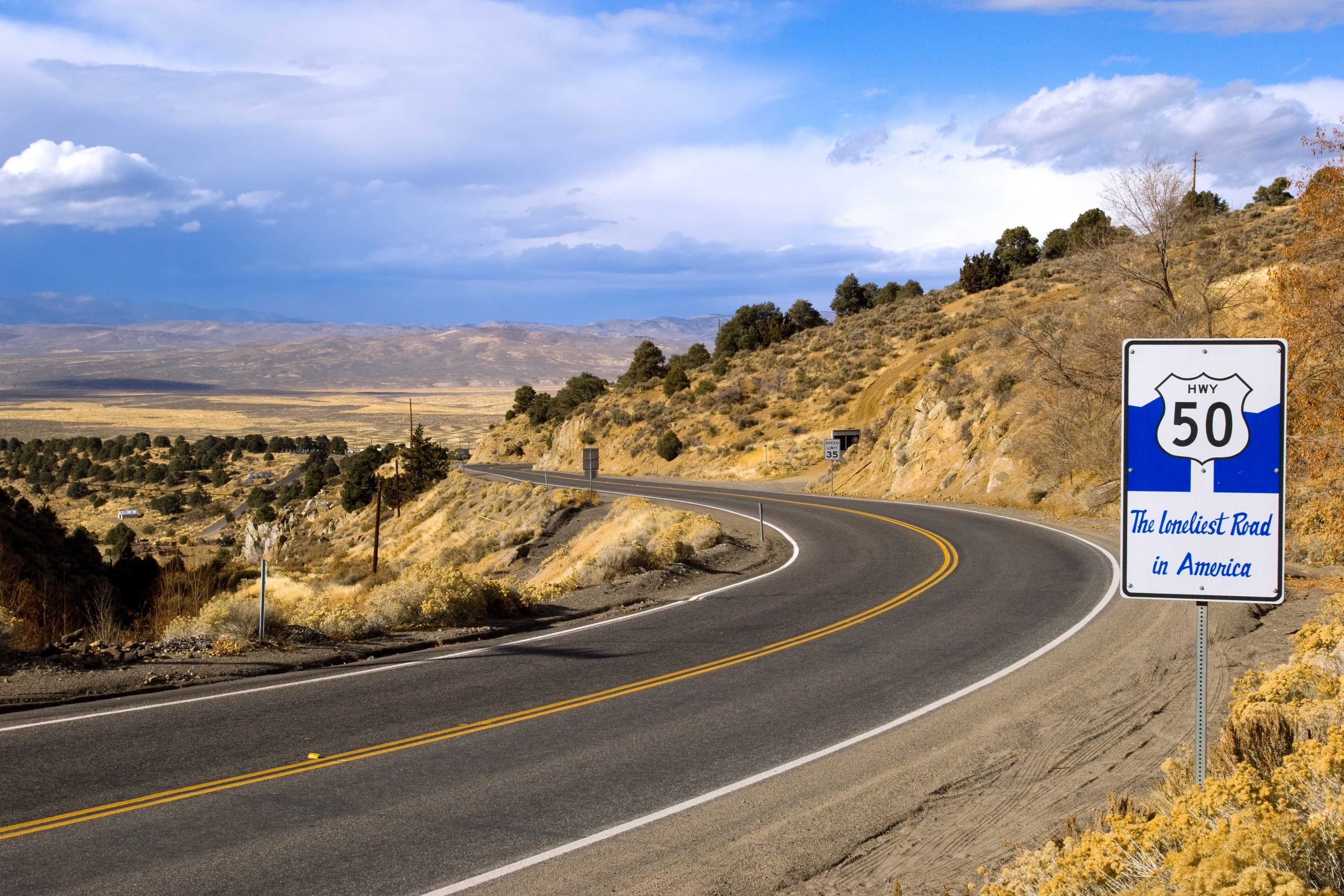 A winding road curves through arid hills under a cloudy sky, with a sign reading "HWY 50: The Loneliest Road in America."