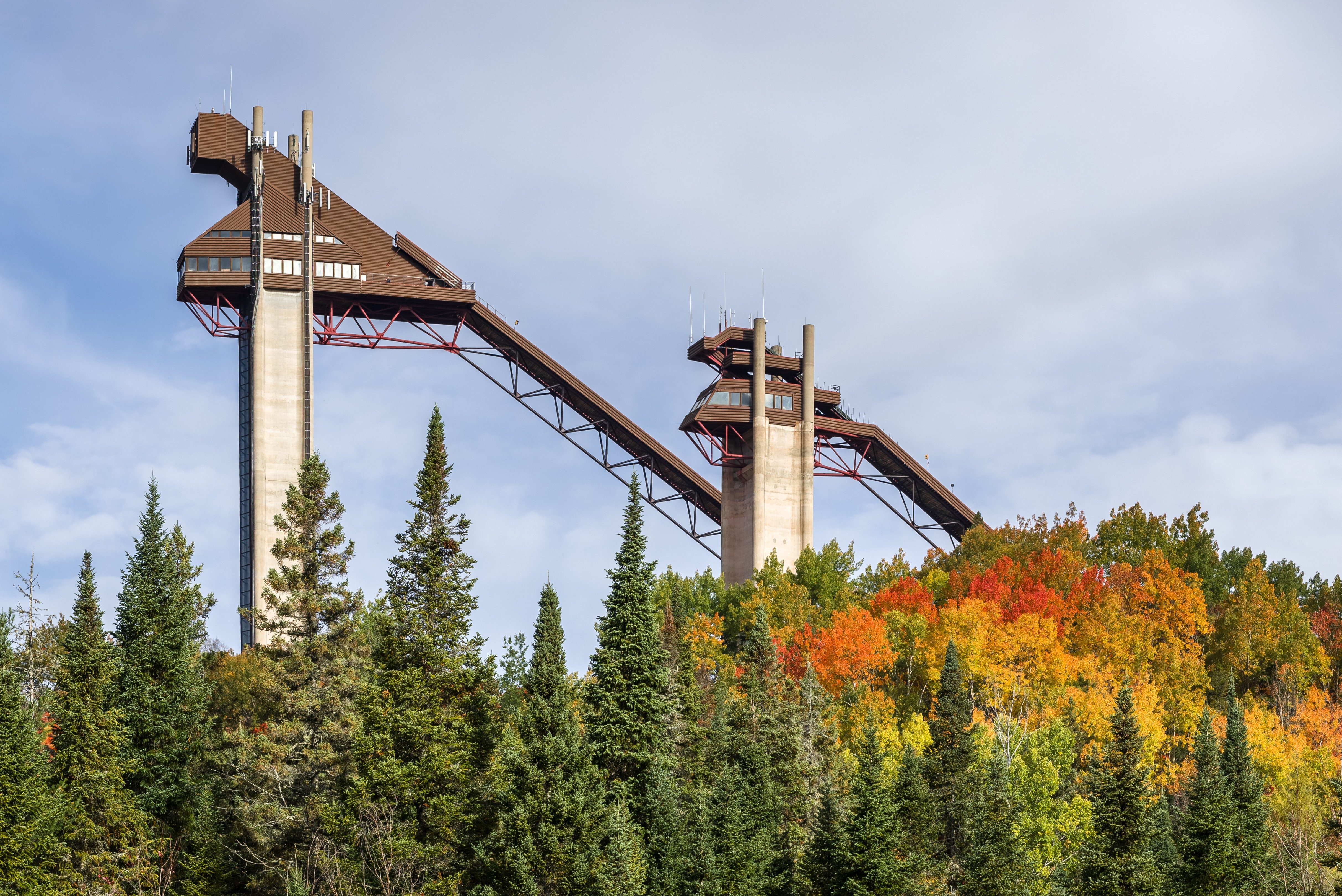 Two large ski jump towers stand amidst colorful autumn trees under a cloudy sky.
