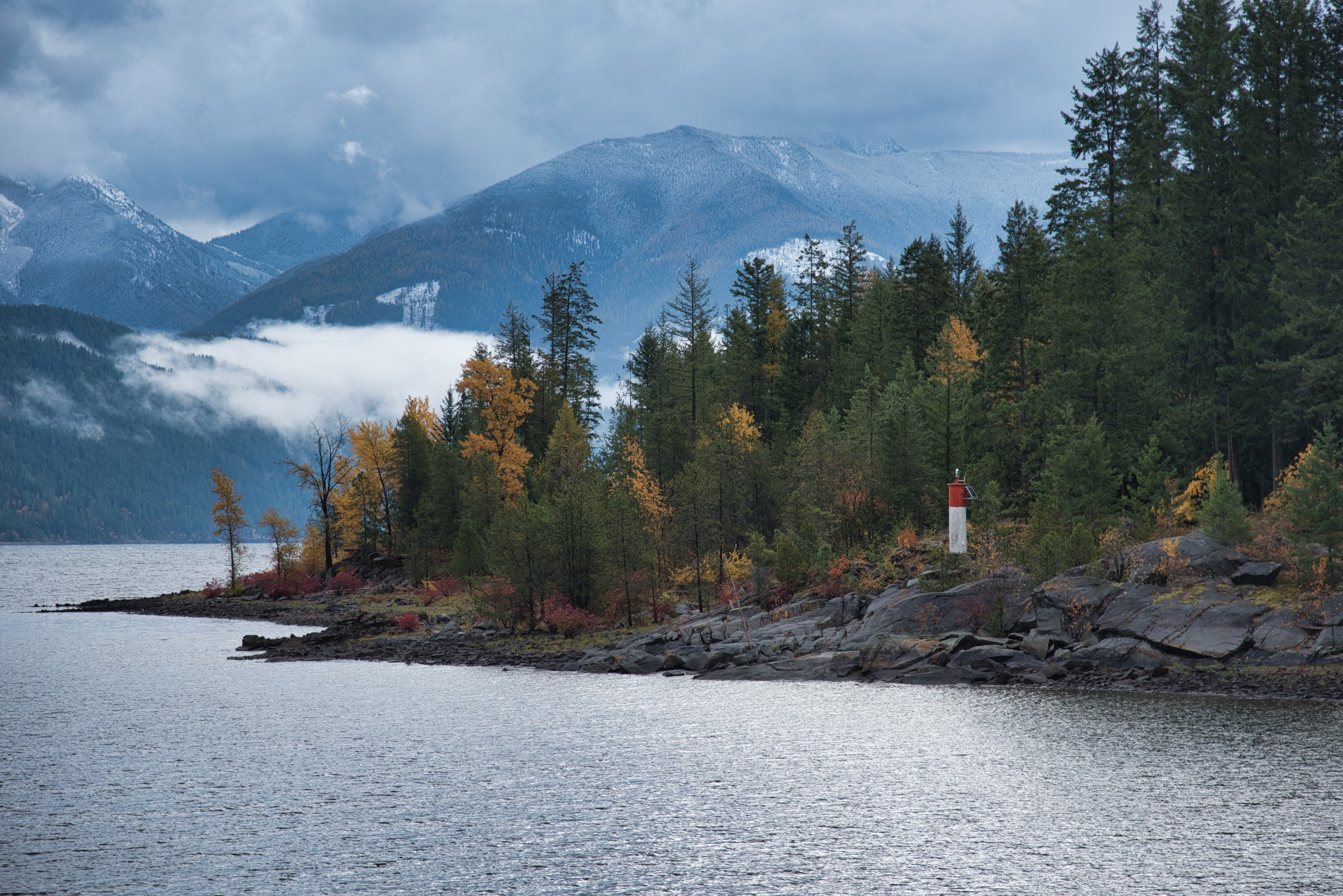 Lighthouse stands quietly by rocky shoreline, surrounded by autumn trees, with misty mountains in the background and a vast, calm lake in front.
