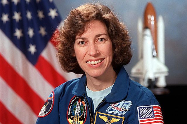 Astronaut smiles in blue uniform adorned with patches, posed in front of American flag and space shuttle model on a blurred background.