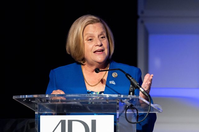 A person speaks at a clear podium, gesturing with a hand. A dark background emphasizes their blue blazer. Text on the podium: "ADL."