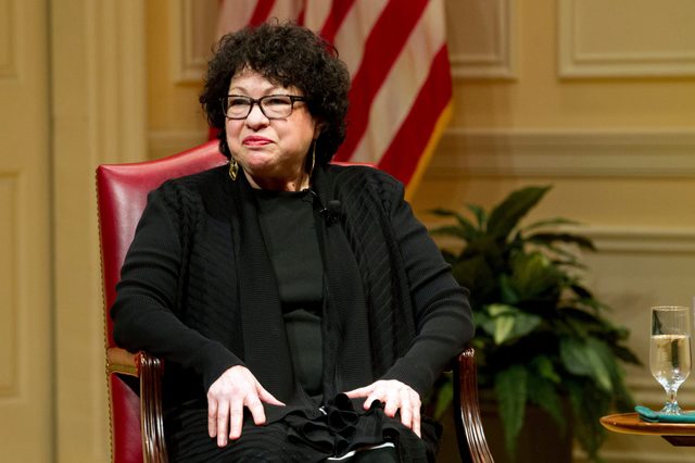 A person sits in a red chair, smiling slightly, in a formal setting with an American flag and plants in the background.