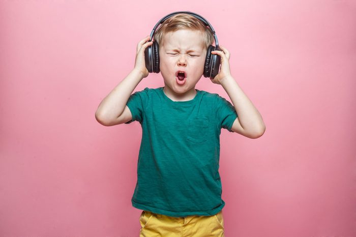 Stylish teen boy listening music in headphones and singing against pink background. School child listening loud music in wireless earphones and dancing. 