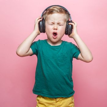 Child wearing headphones, singing enthusiastically against a pink background, wearing a green shirt and yellow shorts.