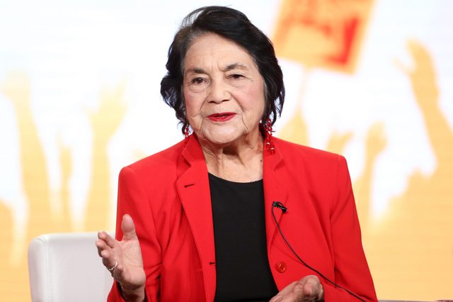 Woman gestures while speaking, seated against a blurred background with raised hands and a sign. She wears a red blazer and matching earrings.