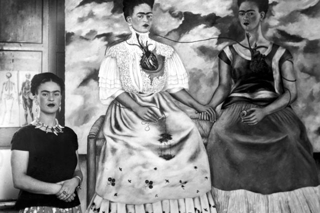 A woman stands beside a painting of two seated women interconnected by veins, against a cloudy sky backdrop and anatomical sketches.