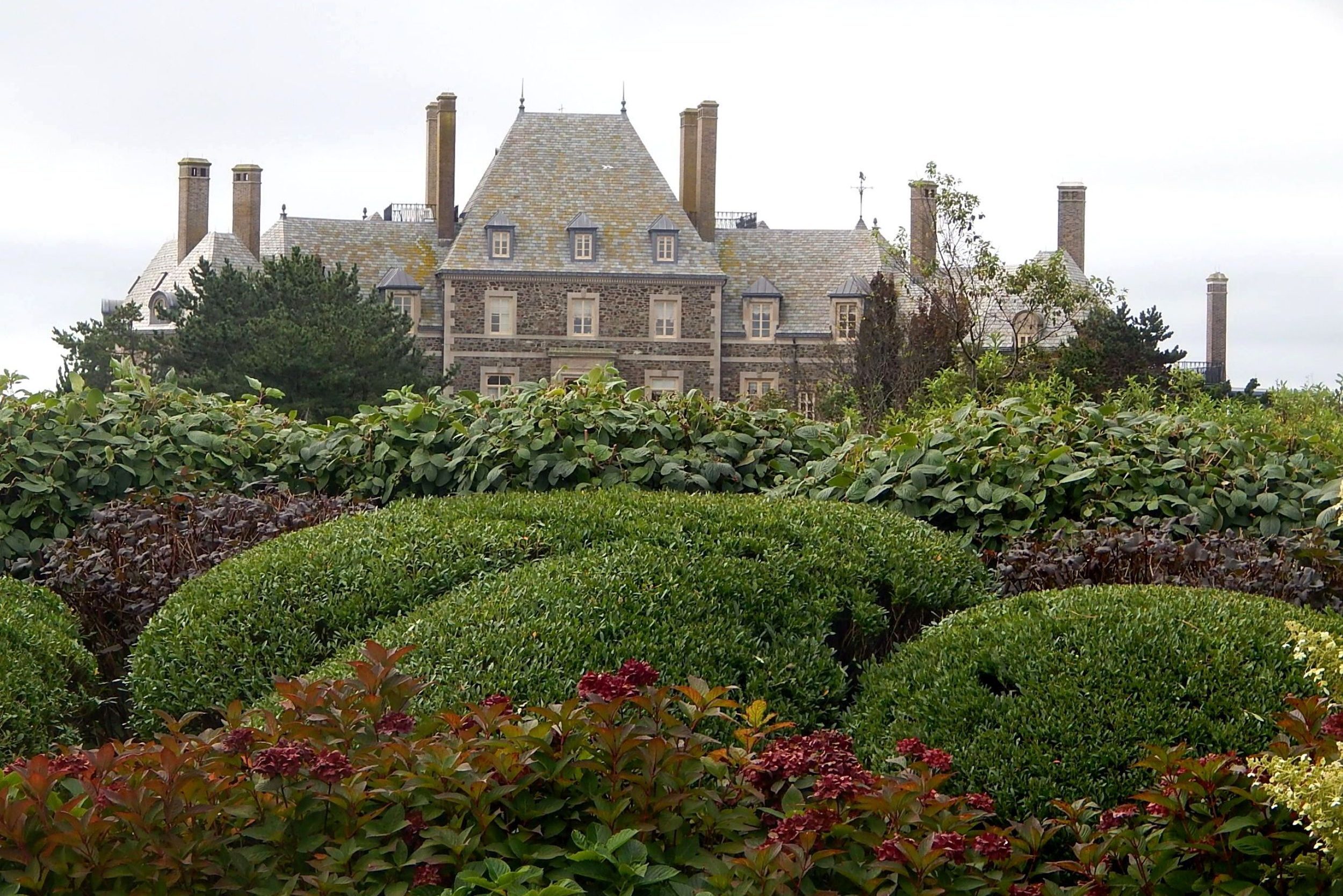 A large mansion stands with chimneys visible above lush green shrubs and trees, under a cloudy sky.